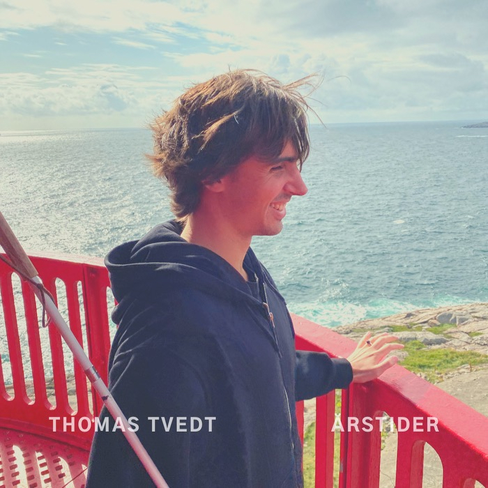 Cover for «Årstider»: Thomas i profil ved et rødt rekkverk, smilende, med havet bak seg og vind i håret.